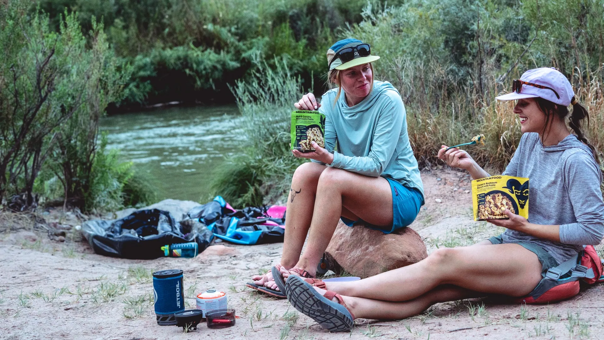 Two campers eating Bighorn meals by a mountain river